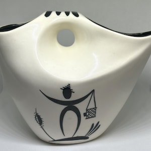 May include: White ceramic vase with a black silhouette of a person carrying a basket. The vase has a unique shape with a hole in the side.