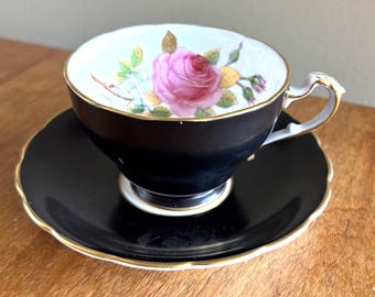 May include: A black teacup and saucer set with a gold trim. The teacup features a pink rose with yellow accents on a white background.