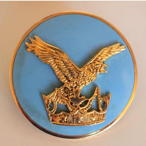 Peut inclure: Un emblème rond bleu clair avec un aigle doré et une couronne. L'aigle est représenté avec les ailes déployées, et la couronne est en bas. L'emblème a une bordure dorée.