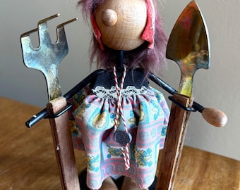 May include: A wooden figurine of a person with pink hair, wearing a floral dress and holding a fork and a trowel. The figurine is standing on a black base.