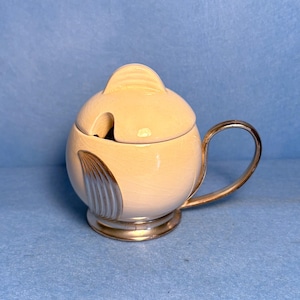 May include: Cream-coloured, egg-shaped sugar bowl with a silver handle and base. The lid has a small, rounded handle and a slot for a spoon. A silver-coloured, stylized wing design adorns the side of the bowl. The bowl is likely made of ceramic.