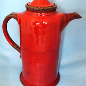 May include: A red ceramic teapot with a brown lid and a brown base. The teapot has a rounded body and a spout on one side.