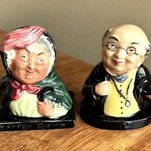 May include: Two ceramic character figurines. The left figure wears a pink bonnet, green shawl, and white blouse, with the text "Sairy Gamp" on the base. The right figure is a bald person with glasses, a black jacket, yellow vest, and a pocket watch chain.