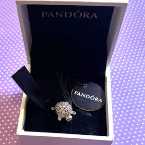 May include: A Pandora charm in a white box. The charm is a small, silver turtle covered in small, sparkling stones. The box is open, revealing a black interior and a tag with the Pandora logo.