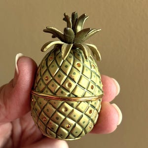 May include: A small, egg-shaped pineapple trinket box with a matte olive green finish and a textured diamond pattern. The surface is accented with small, embedded amber-coloured crystals. It features a hinged lid and a metallic green leaf top.