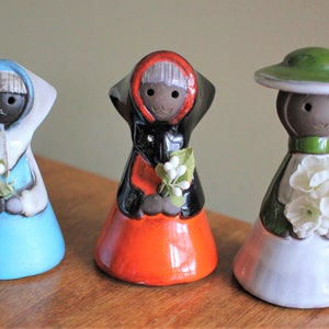 May include: Three ceramic figurines of women wearing different colored dresses and hats, each holding a bouquet of white flowers. The woman on the left is wearing a blue dress and a white hat, the woman in the middle is wearing an orange dress and a red hat, and the woman on the right is wearing a white dress and a green hat.