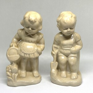 May include: Two ceramic figurines of children. The girl is holding a pail and wearing a dress. The boy is holding a shovel and wearing overalls. Both figurines are painted in a cream color.