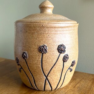 May include: A brown ceramic jar with a lid, decorated with black flower designs and brown raised flower buds.