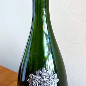 May include: A green glass bottle with a silver emblem and base. The bottle has a decorative silver emblem in the center and a silver band at the base with text. The bottle is on a wooden surface.