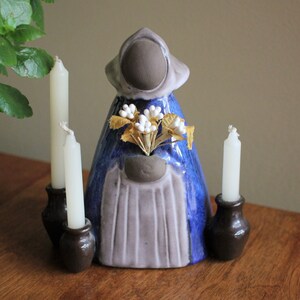 May include: A ceramic candle holder featuring a woman in a blue and grey robe holding a bouquet of white flowers. The holder has two candle holders on either side.
