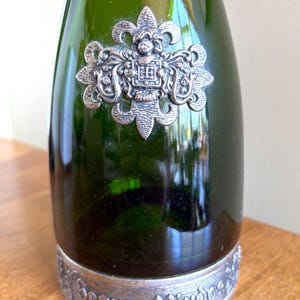 May include: Green glass bottle with a silver-colored decorative emblem and base. The emblem features an ornate design with a crest. The base of the bottle has the words "Segura Viudas" in a decorative font.