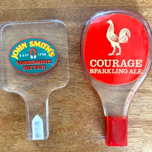 May include: Two vintage beer tap handles. One is clear with a John Smith's Yorkshire Bitter logo. The other is red with a white rooster and the words Courage Sparkling Ale.