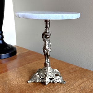 May include: A small decorative table with a round, white marble top. The base is a detailed, silver-toned sculpture of a woman. The base has an ornate, floral-like design. The table is on a wooden surface.