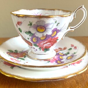 May include: A white teacup, saucer, and side plate set with a floral design in purple, red, and yellow. The edges are trimmed with gold. The teacup has a curved handle.