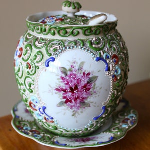 May include: A white porcelain jar with a green, blue and red floral design. The jar has a lid and a matching saucer. The jar is decorated with raised floral details and a spoon.