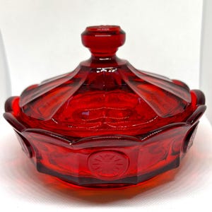 Peut inclure: Un plat à bonbons en verre rouge avec un couvercle. Le plat a un bord festonné et un motif en étoile sur le fond.