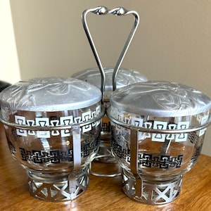 May include: A set of three silver and glass condiment jars with black and white geometric designs. The jars have lids and are attached to a silver wire rack with a handle.