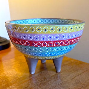 May include: A colorful, hand-painted ceramic bowl with three gray legs. The bowl features horizontal bands of yellow, pink, red, and blue, each adorned with a pattern of white and blue dots. A decorative piece for the home.