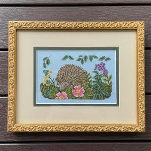 May include: A cross-stitch embroidery featuring a brown hedgehog surrounded by green foliage, pink flowers, and yellow blossoms against a light blue background, encased in a rectangular gold frame with a floral relief pattern and a cream mat.