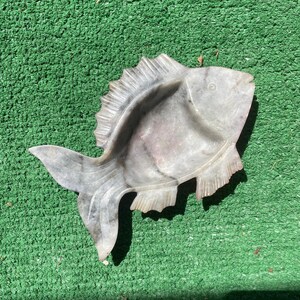 May include: A carved stone dish shaped like a fish with a smooth, polished surface. The object features a light gray and white marbled pattern with subtle veining throughout. It has a recessed center, a defined tail, and etched fin details.