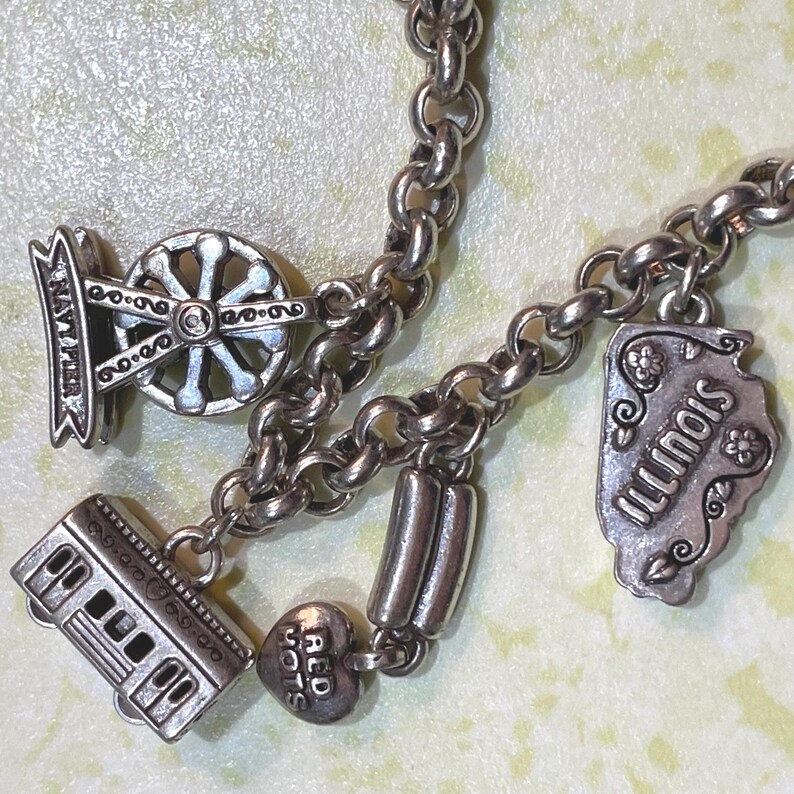 May include: Silver charm bracelet with multiple charms. Charms include a Navy Pier emblem, a train, a heart with "Red Hots" text, a state of Illinois charm, and two cylindrical charms. The bracelet is on a light yellow surface.