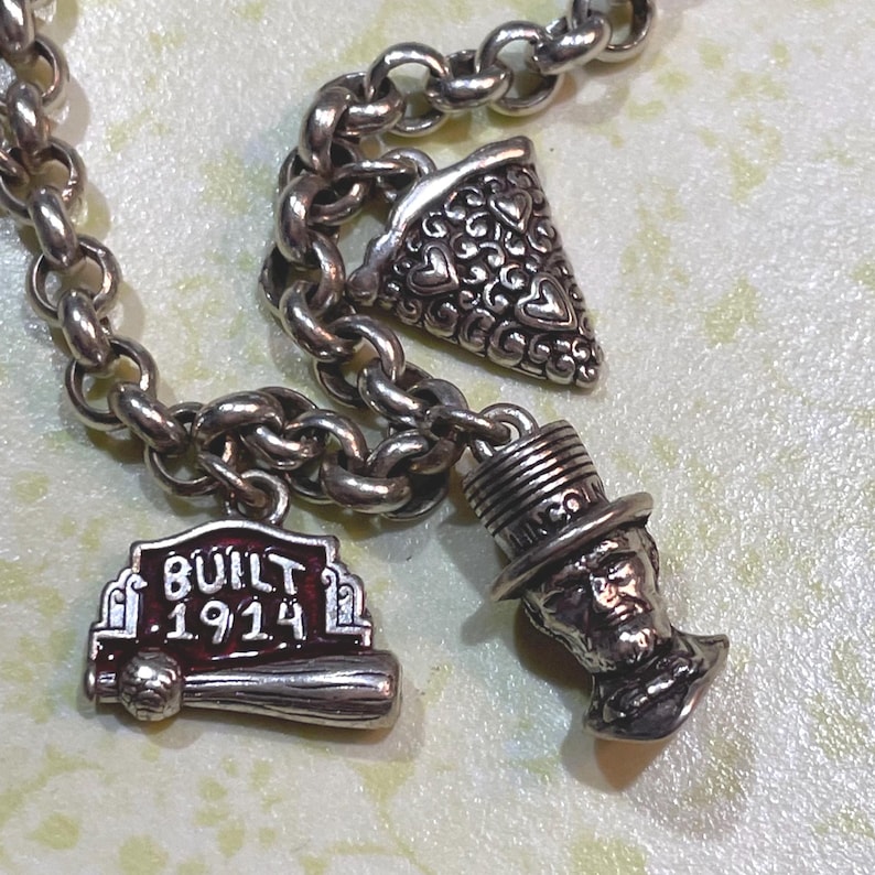 May include: A silver charm bracelet with three detailed charms. One charm is a baseball bat with the text "BUILT 1914". Another charm is a slice of pizza with heart-shaped cutouts. The third charm is a bust of a man wearing a top hat.