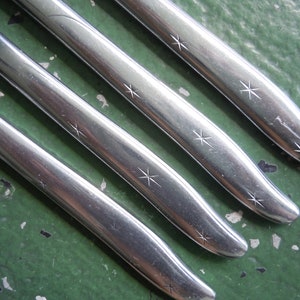 May include: Close-up of four stainless steel utensils, possibly forks or knives, arranged diagonally. Each utensil has a polished finish and is adorned with a series of starburst designs. The background is a mottled green surface.