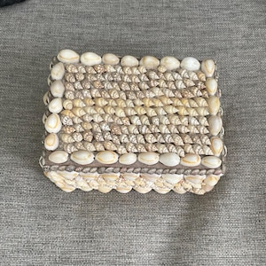 May include: A rectangular box covered in various types of seashells. The lid features rows of small, tan, spiral-shaped shells bordered by larger, smooth, white and cream-colored shells. The sides are also decorated with layers of small shells.