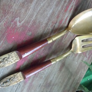 May include: A brass serving spoon and a three-tined brass serving fork with dark brown wooden handles. The ends of the handles feature decorative brass caps with embossed figures. The metal surfaces are polished and have a warm golden tone.