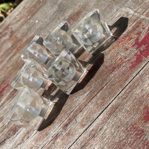 May include: A collection of six clear, square-shaped cabinet knobs made of transparent material. Each knob features a smooth, cubic exterior with a circular recessed center and a metal screw attachment visible on the underside of the pieces.