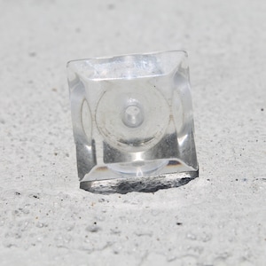 May include: A clear, square-shaped cabinet knob with a central screw hole. The knob is made of transparent material, reflecting light and set against a textured, light-colored surface. The design is simple and modern.