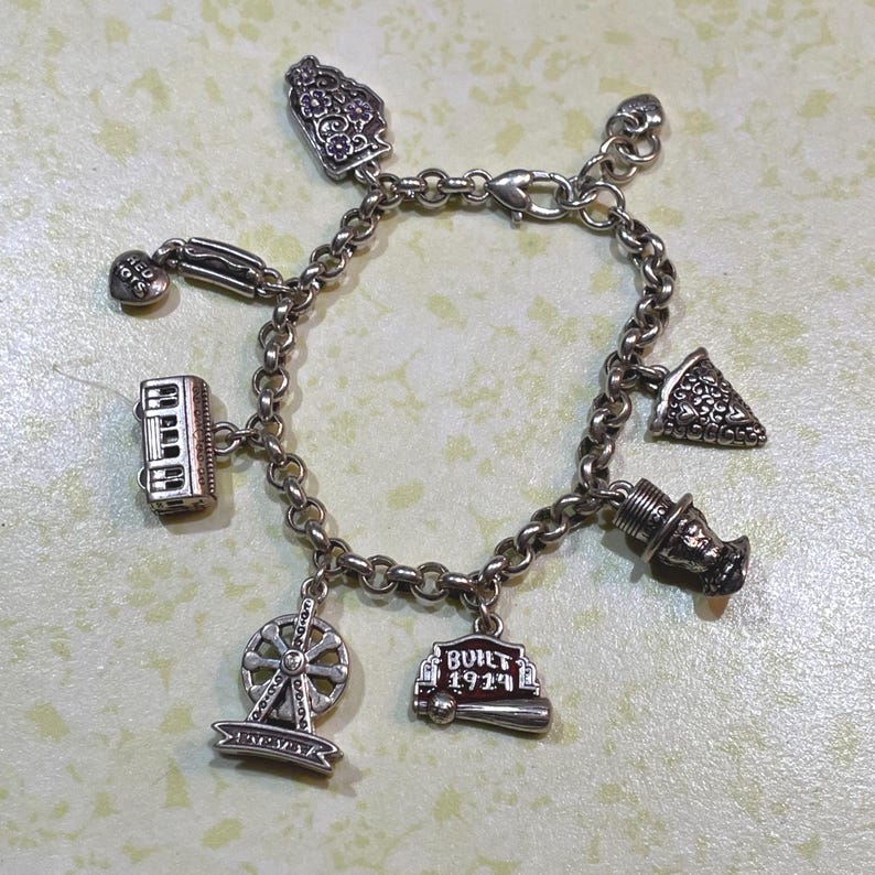 May include: Silver charm bracelet featuring various charms, including a Ferris wheel, a building with "BUILT 1924" text, and a pizza slice. The bracelet has a lobster clasp closure and is displayed on a light green surface.