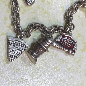 May include: Silver charm bracelet with multiple charms. Charms include a pizza slice with "Chicago Pizza" text, a top hat, and a "Wrigley Field" charm. The bracelet is on a light green surface.