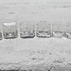 May include: Six clear, square cabinet knobs with a circular design in the center. The knobs are made of a transparent material and are arranged in a row. The knobs are sitting on a white surface.