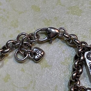 May include: Close-up of a silver-toned necklace chain with a heart-shaped clasp and a small heart charm. The chain links are round and connected. The charm has an ornate design.