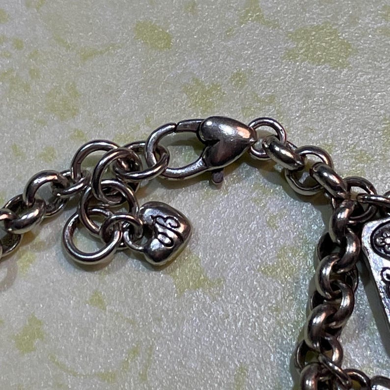 May include: Close-up of a silver-toned necklace chain with a heart-shaped clasp and a small heart charm. The chain links are round and connected. The charm has an ornate design.