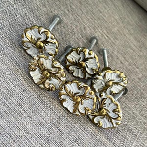 May include: A set of five round cabinet knobs featuring a white base with raised gold-toned floral and leaf-like scrollwork patterns. Each knob has a central circular detail and a silver-toned metal bolt extending from the back for installation.