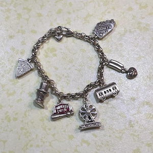 May include: Silver charm bracelet featuring various charms, including a slice of pizza, a Ferris wheel, a train, and a baseball bat with "Wrigley Field" text. The bracelet also has charms with the text "Illinois" and "Old Town".