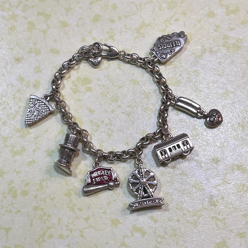 May include: Silver charm bracelet featuring various charms, including a slice of pizza, a Ferris wheel, a train, and a baseball bat with "Wrigley Field" text. The bracelet also has charms with the text "Illinois" and "Old Town".