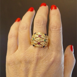 Puede incluir: Un anillo dorado con un diseño texturizado, engastado con pequeñas piedras preciosas rojas, verdes y azules. El anillo se lleva en una mano con esmalte de uñas rojo.