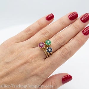 May include: A gold ring featuring three flower-shaped clusters, each set with a central diamond surrounded by small rubies, emeralds, or sapphires. The band has a twisted rope texture, and the ring is worn on a finger with red nail polish.