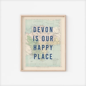 May include: A framed print displaying a vintage map of Devon, England. The map is in cream, green, and yellow tones, with the navy text "Devon is our happy place". The print is encased in a light wooden frame.