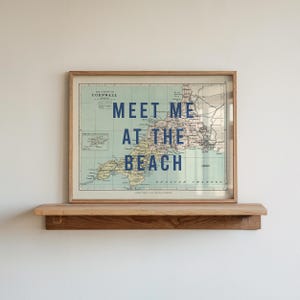 May include: A framed map of Cornwall with the text "MEET ME AT THE BEACH" in navy blue. The map is in a wooden frame and sits above a wooden shelf, mounted on a white wall.