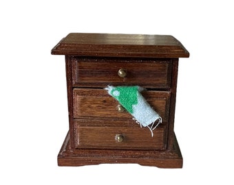 May include: A small wooden dresser with four drawers. The top drawer is open and a green and white fabric is hanging out of it.