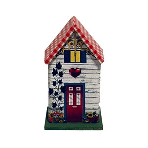 May include: A decorative miniature house with a red and white checkered roof, a red door, and a heart-shaped accent. The house has a white exterior with a vine design and sits on a green base. The window has a yellow and blue design.