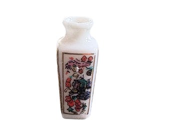 May include: A small, white porcelain vase with a rectangular shape and a narrow neck. The vase is decorated with a colorful floral and bird design, featuring red, green, and gold accents. The vase is likely a decorative item.