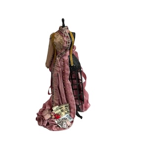May include: A miniature dress form displays a dusty pink gown with a long train, featuring a black cage skirt underneath. The bodice is adorned with a yellow measuring tape trim, small buttons, and miniature paper ephemera at the hemline.