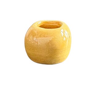 May include: A small, round, yellow ceramic vase with a glossy finish. The vase has a wide opening and a slightly textured surface. The color is a warm, golden yellow, and the overall shape is organic and simple.