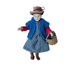 May include: A small doll wearing a blue and white checkered dress, a blue cardigan, and a red sweater. The doll is wearing a brown hat with a red ribbon and is holding a brown bucket.