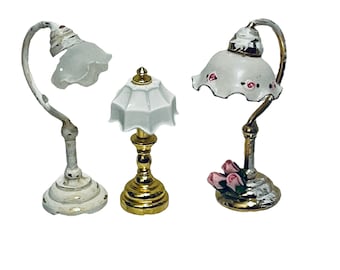 May include: Three miniature table lamps with white shades. The lamp on the left has a white base, the middle lamp has a gold base, and the lamp on the right has a silver base with pink roses.